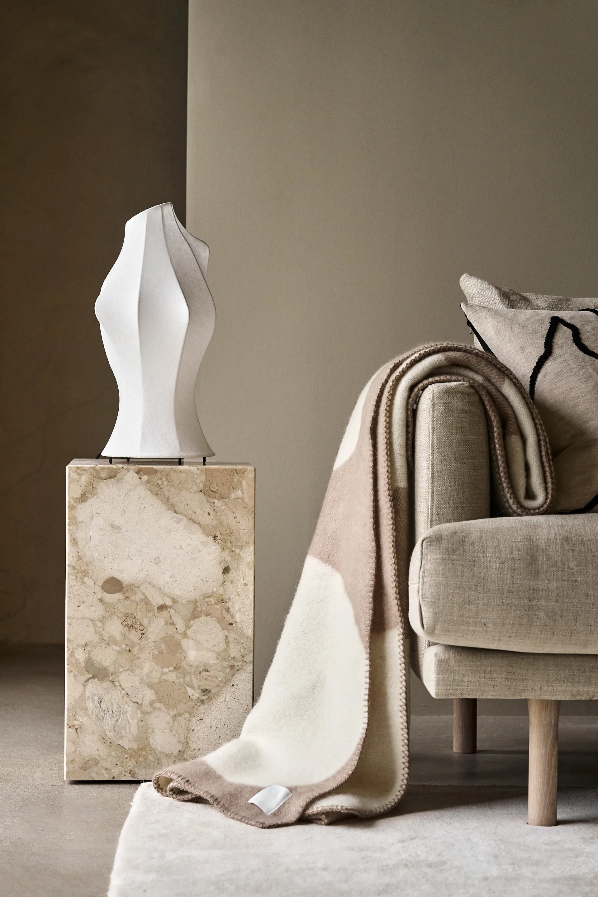 Vit skulptural lampa på marmor piedestal, bredvid en ljus beige soffa med en vikad gräddvit och brun pläd och en mönstrad kudde.