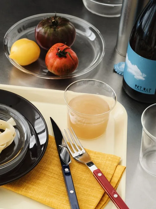En beige bricka med pasta, juice, bestick, citroner, tomater, salt, peppar och en vinflaska på ett metallbord.
