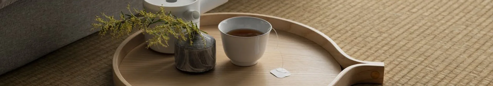 En ljus träbricka med en vit tekopp med tepåse, en grå vas med en gulgrön gren, och en vit behållare på en texturerad matta.