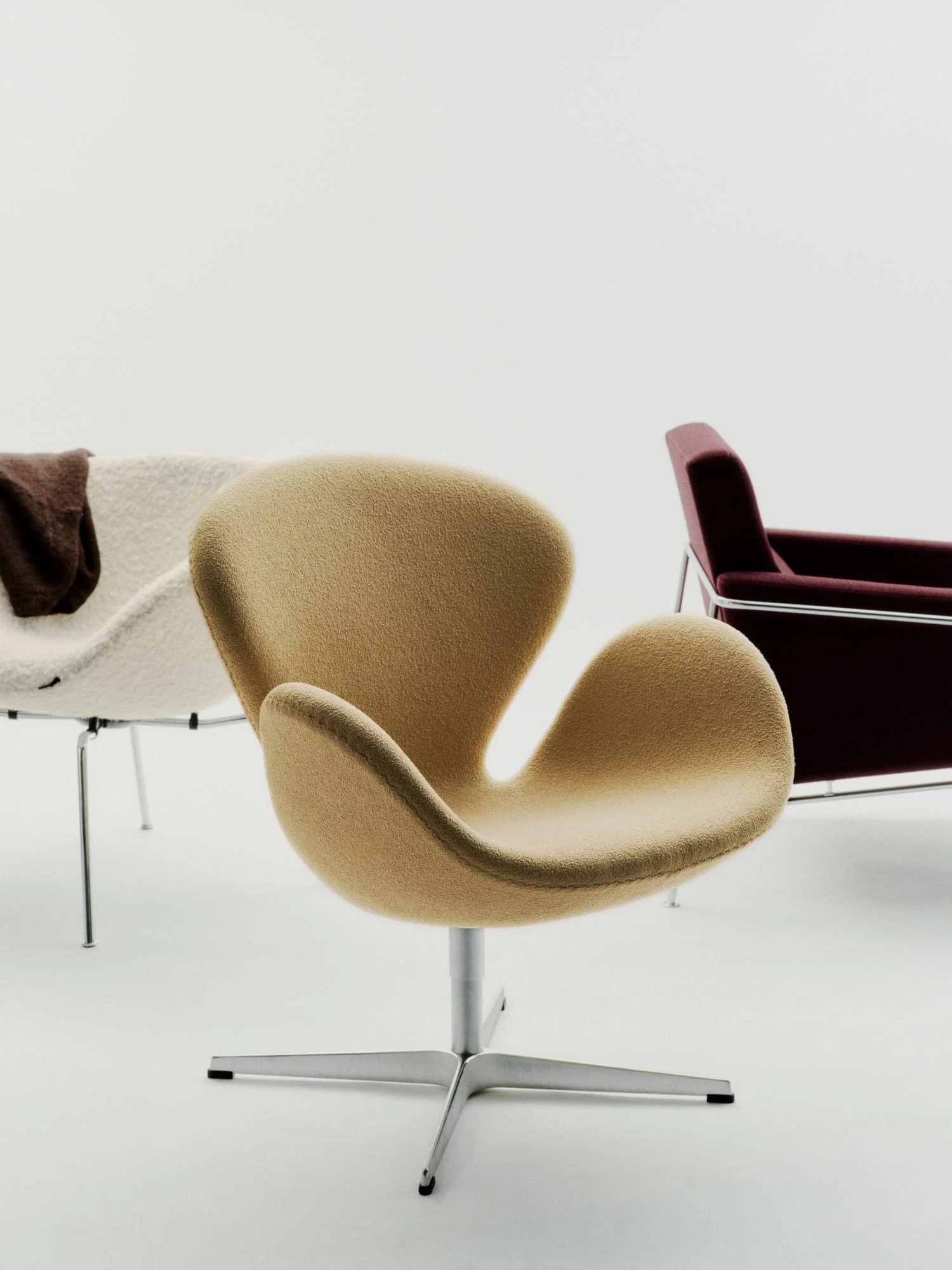 Tre stolar på vit bakgrund. Fram, en beige, böjd stol i texturerat tyg. Bakom, en krämfärgad stol och en röd fåtölj.