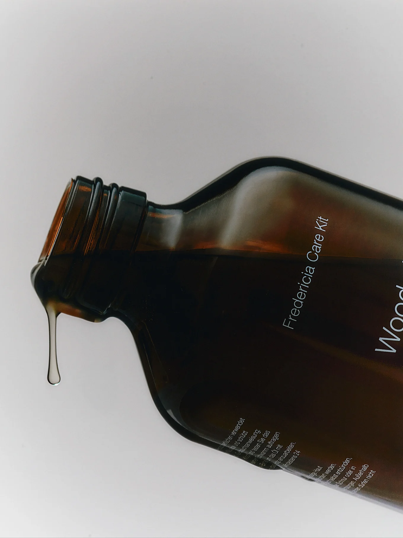 En lutande, mörk, bärnstensfärgad flaska från Fredericia Care Kit, med en droppe olja som droppar från flaskhalsen.