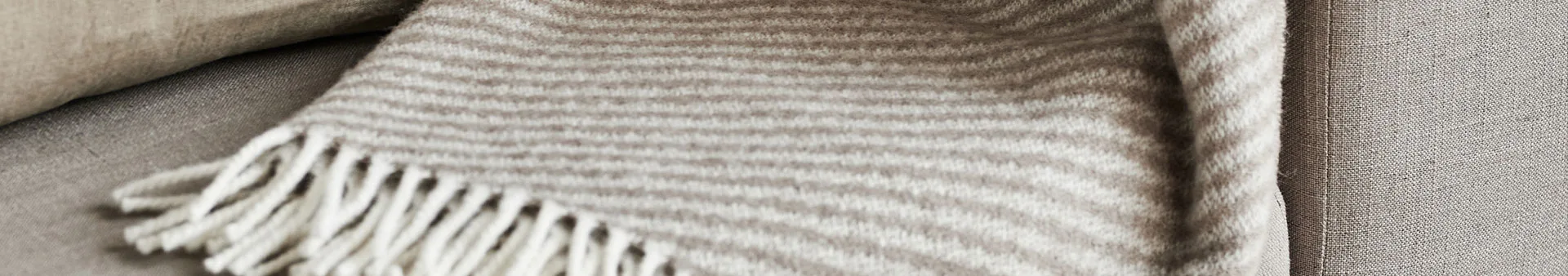 En randig filt med fransar i beige och offwhite, liggande över en ljusbrun textil yta.