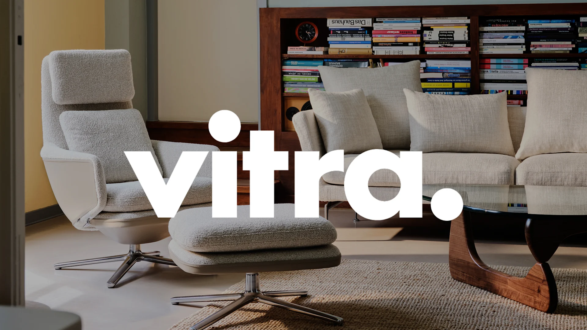 Vitra.-logotyp över modern inredning med ljus textil fåtölj, fotpall och soffa, samt bokhyllor.
