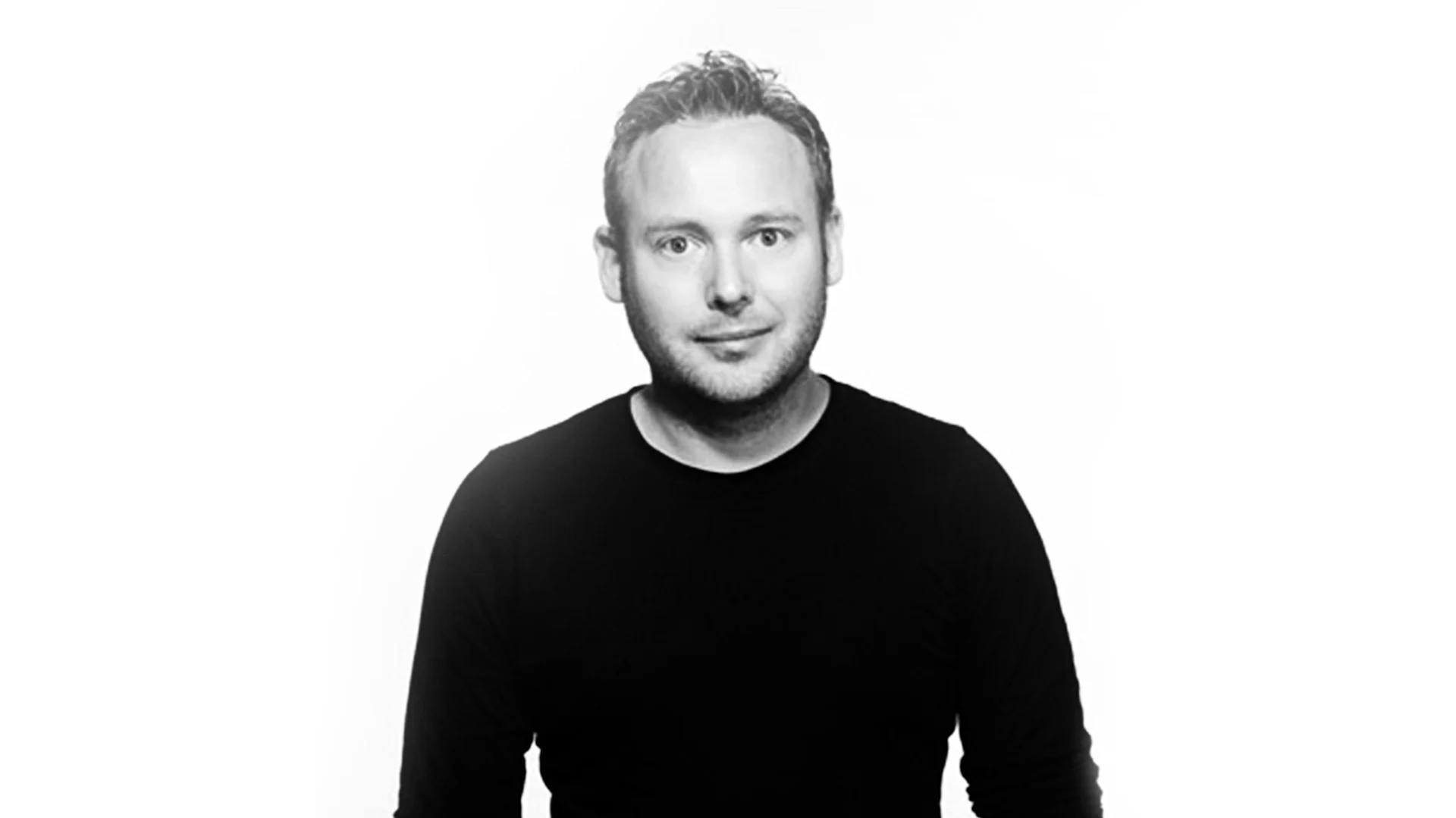 Svartvitt porträtt av en leende man med kort hår och skägg, iklädd en mörk långärmad tröja.